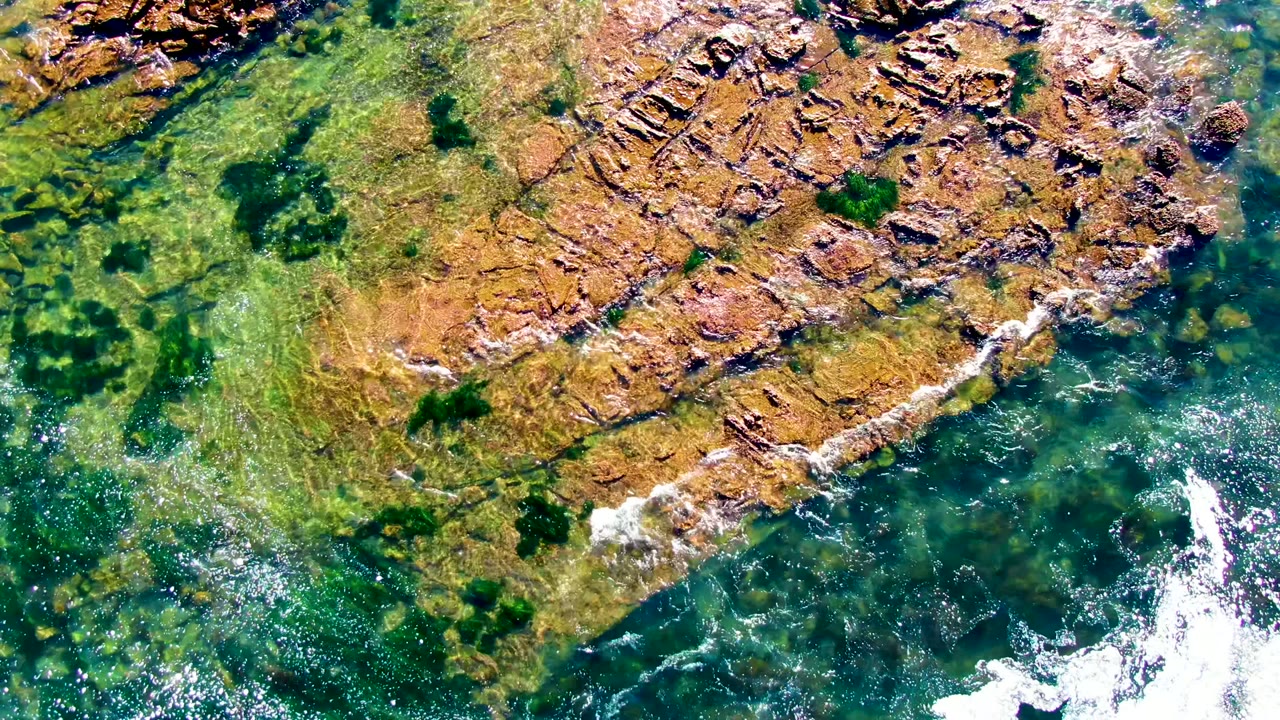 Aerial view in La Bocana, Baja California Sur, showing clear green-blue water, brown rock shelves, and white foam at the start of the clip, with no visible people.