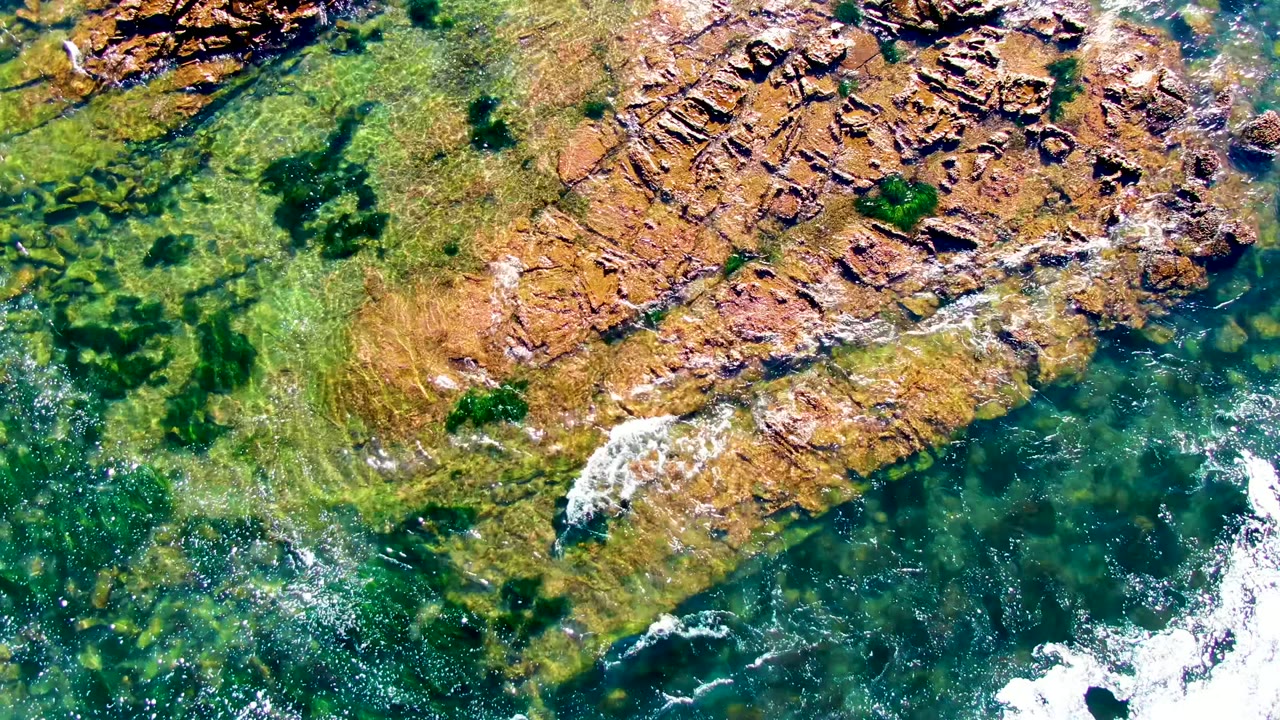 Aerial view in La Bocana, Baja California Sur, showing reddish-brown shoreline rocks, transparent green water, and a white foam patch near the lower right, with no visible people.