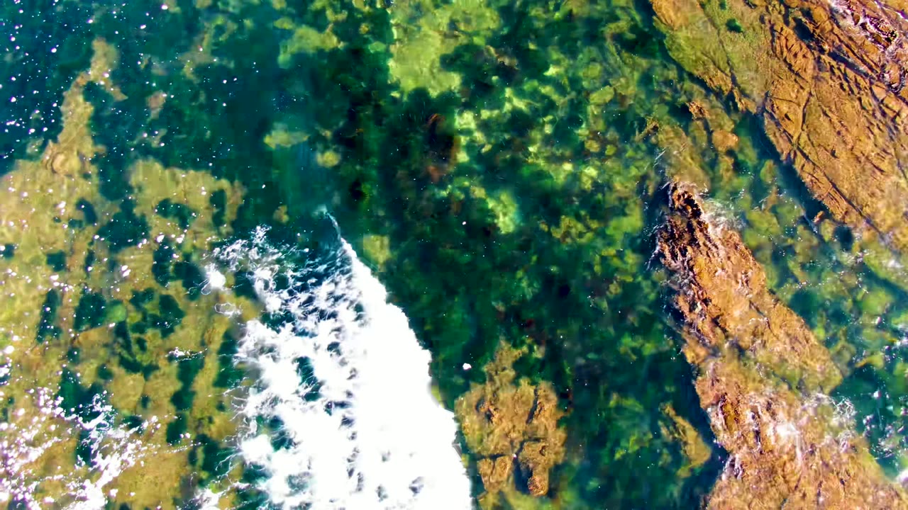 Top-down view of clear green-blue water over brown rocky seabed in La Bocana, Baja California Sur, with a band of white foam entering from the lower left and no visible people.