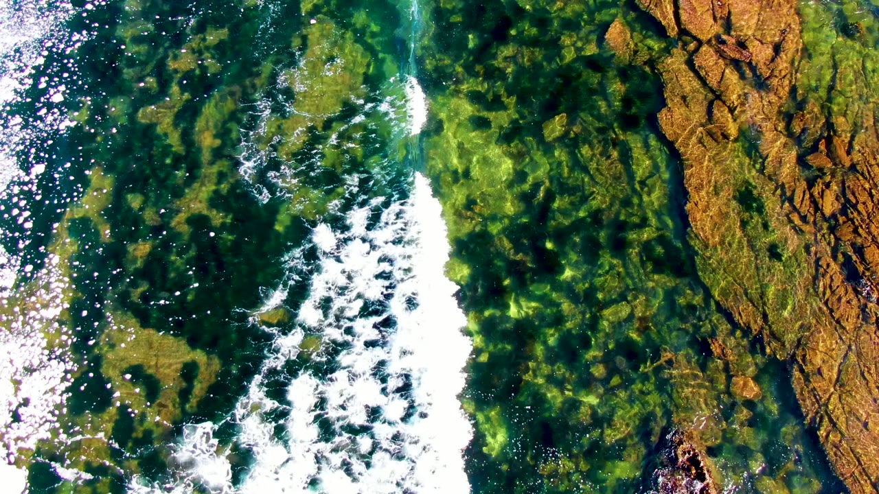 Overhead view in La Bocana, Baja California Sur, showing white surf foam moving across green shallow water and brown rock shelves, with no visible people.