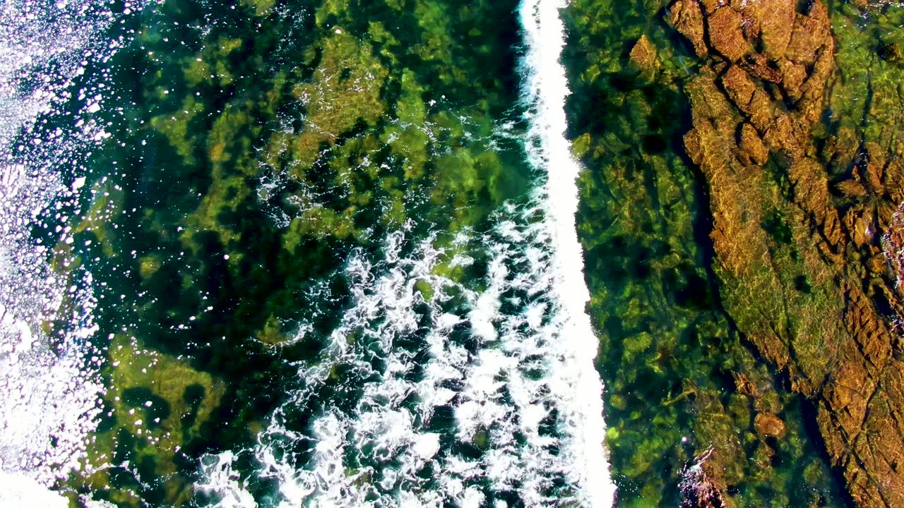 A vertical band of white foam divides clear green water from exposed brown rock texture in La Bocana, Baja California Sur, with no visible people.