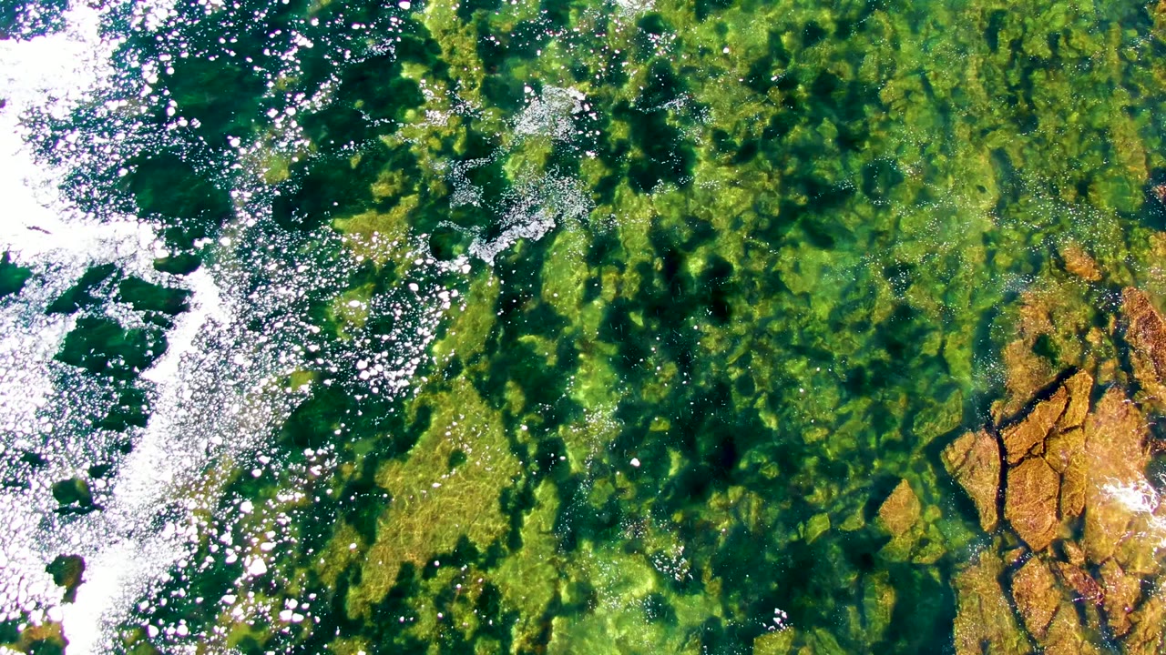 Overhead frame from La Bocana, Baja California Sur showing clear green shallow water, white surf on the left, tan rocks at the lower right, and no visible people.