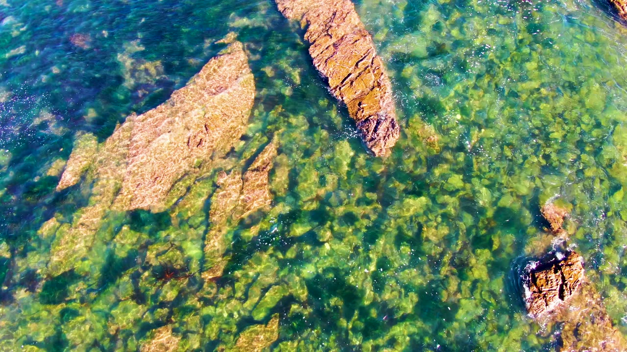 Overhead frame of clear green-blue shallow water over tan rocks in La Bocana, Baja California Sur, with no visible people.