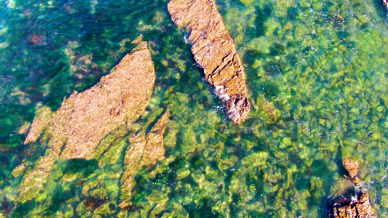 Overhead La Bocana frame with a tan rock shelf left of center, a long rock ridge near the upper right, clear green water, and no visible people.