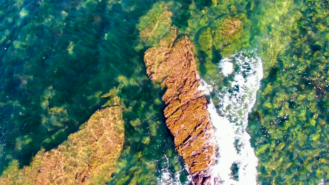 Overhead frame from La Bocana, Baja California Sur showing clear green ocean water, tan submerged rocks, a longer exposed rock band, white foam along its edge, and no visible people.
