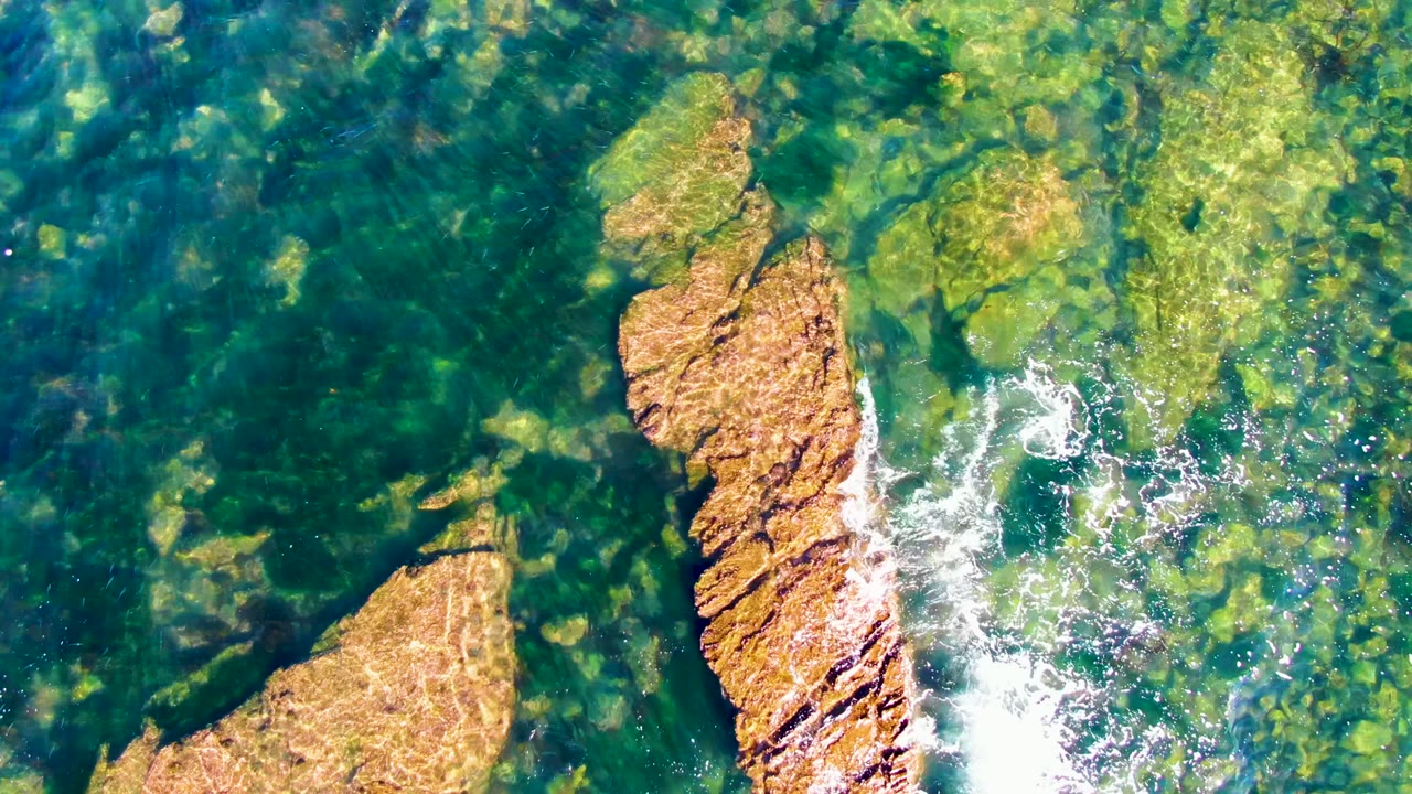 La Bocana overhead frame showing a diagonal tan rock ridge, mottled underwater stones, bright white foam beside the ridge, and no visible people.