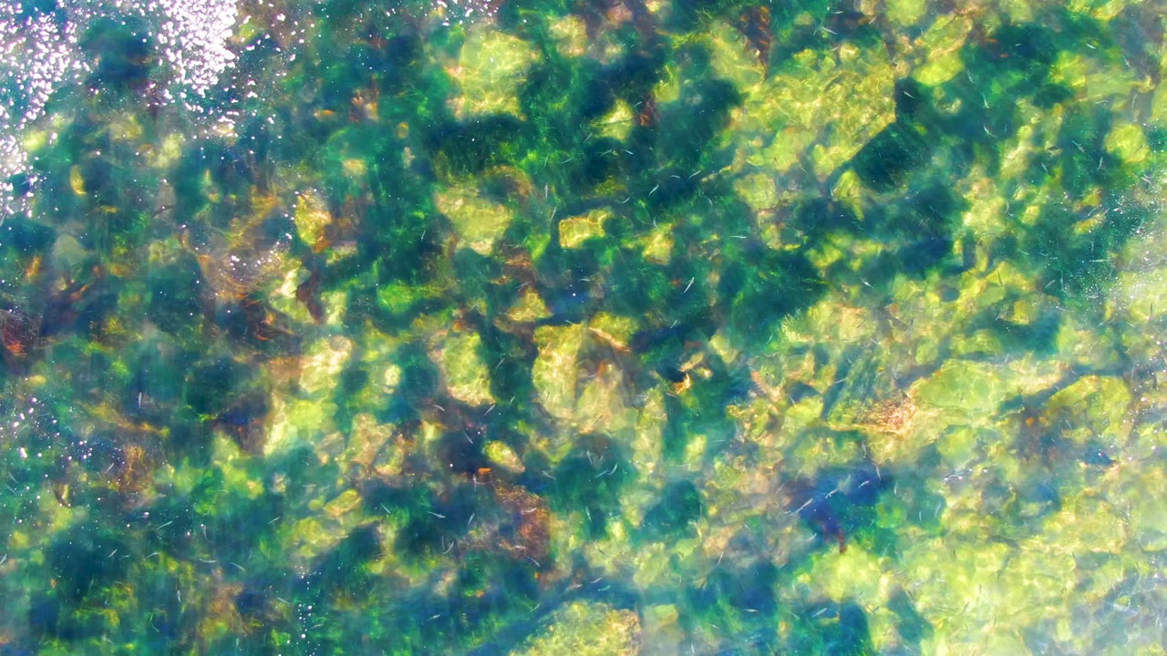 Clear shallow water in La Bocana, Baja California Sur shows green-blue submerged rocks and bright surface sparkle with no visible people.