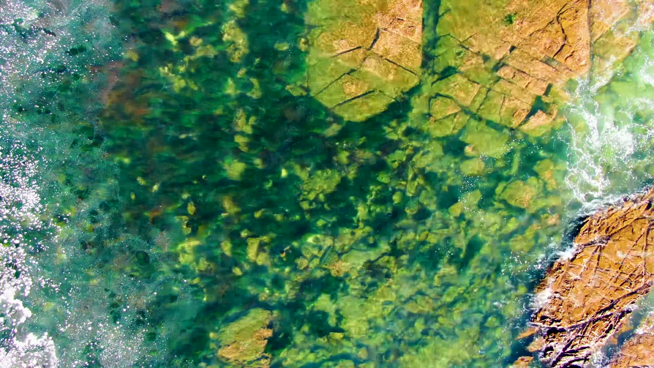 Overhead view of clear turquoise water over tan rocks at La Bocana, Baja California Sur, with small white foam patches and no visible people.