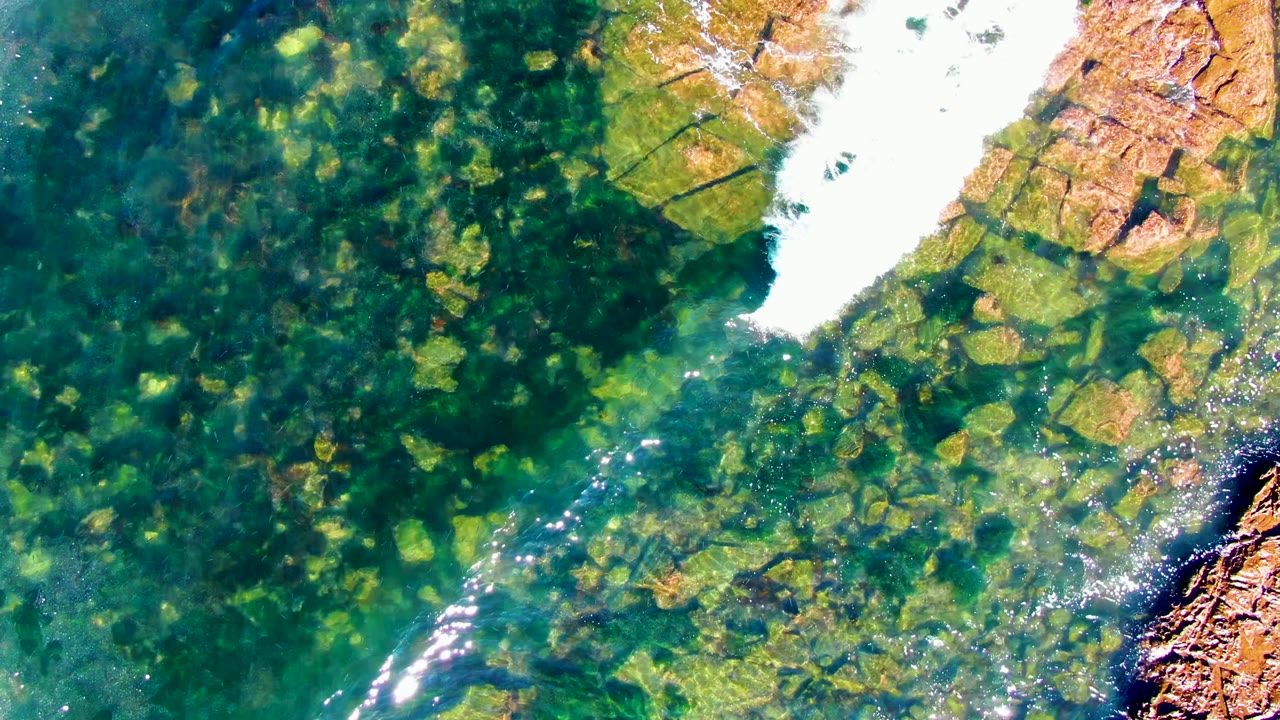 Aerial overhead frame of green-blue clear water over a rocky bed in La Bocana, Baja California Sur, with bright white foam entering from the upper right and no visible people.