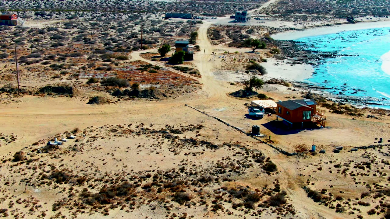 Aerial frame over La Bocana shows a white car beside a small wood-colored building, a pale dirt road, scrubby desert ground, bright blue water, and no visible people.