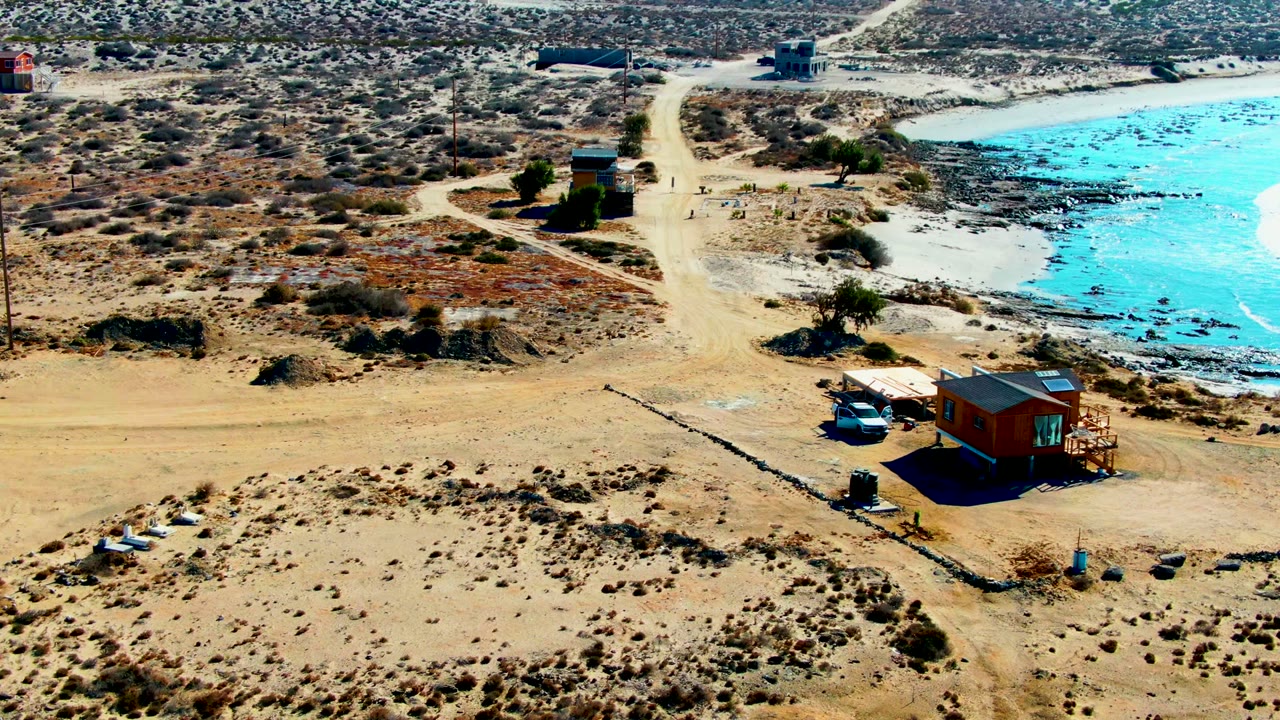 White car and small building beside a dirt road near blue coastal water.