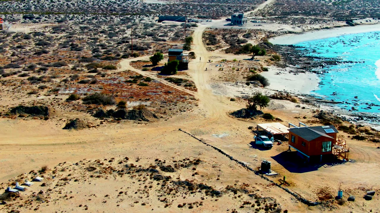 La Bocana aerial frame shows the small wood-colored structure and parked or slow-moving white car near the lower right, with dry scrubland, a dirt road, blue water, and no visible people.
