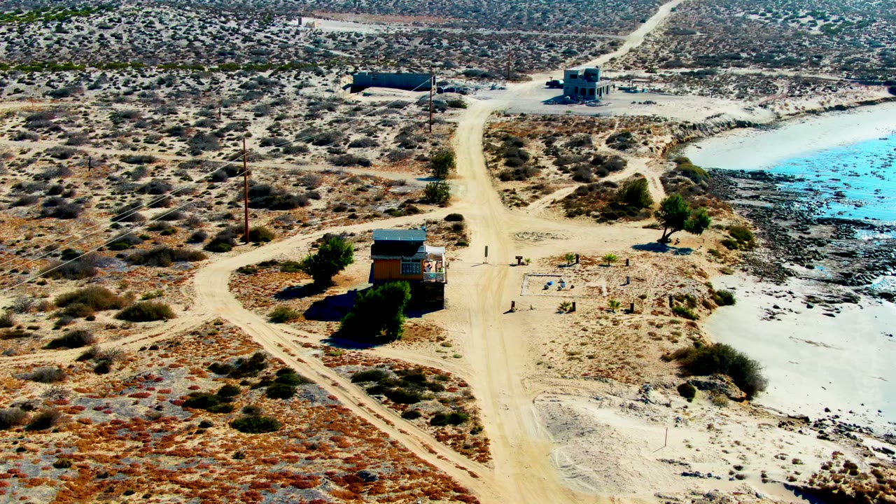 Elevated drone view in La Bocana, Baja California Sur, showing a small cabin near intersecting dirt roads, scrub desert, and a blue-green reservoir, with no visible people.