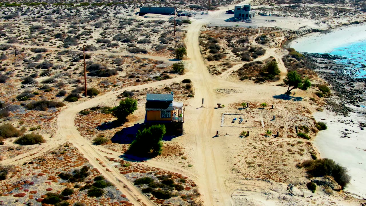 Mid-clip aerial view in La Bocana, Baja California Sur, showing the cabin near the lower right, dirt tracks crossing the scrub, utility poles, and bright reservoir water beyond, with no visible people.