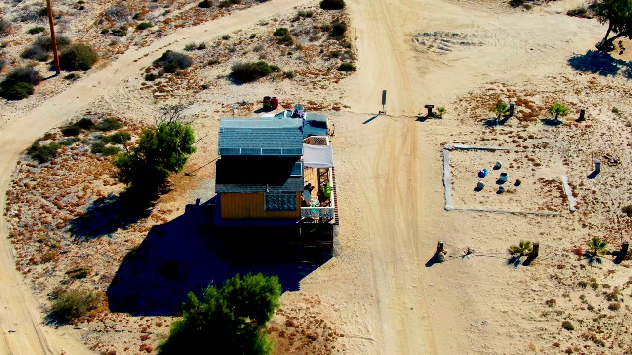 Aerial frame in La Bocana, Baja California Sur showing a white truck on sandy ground near a small house with rooftop solar panels, sparse shrubs, and no visible people.