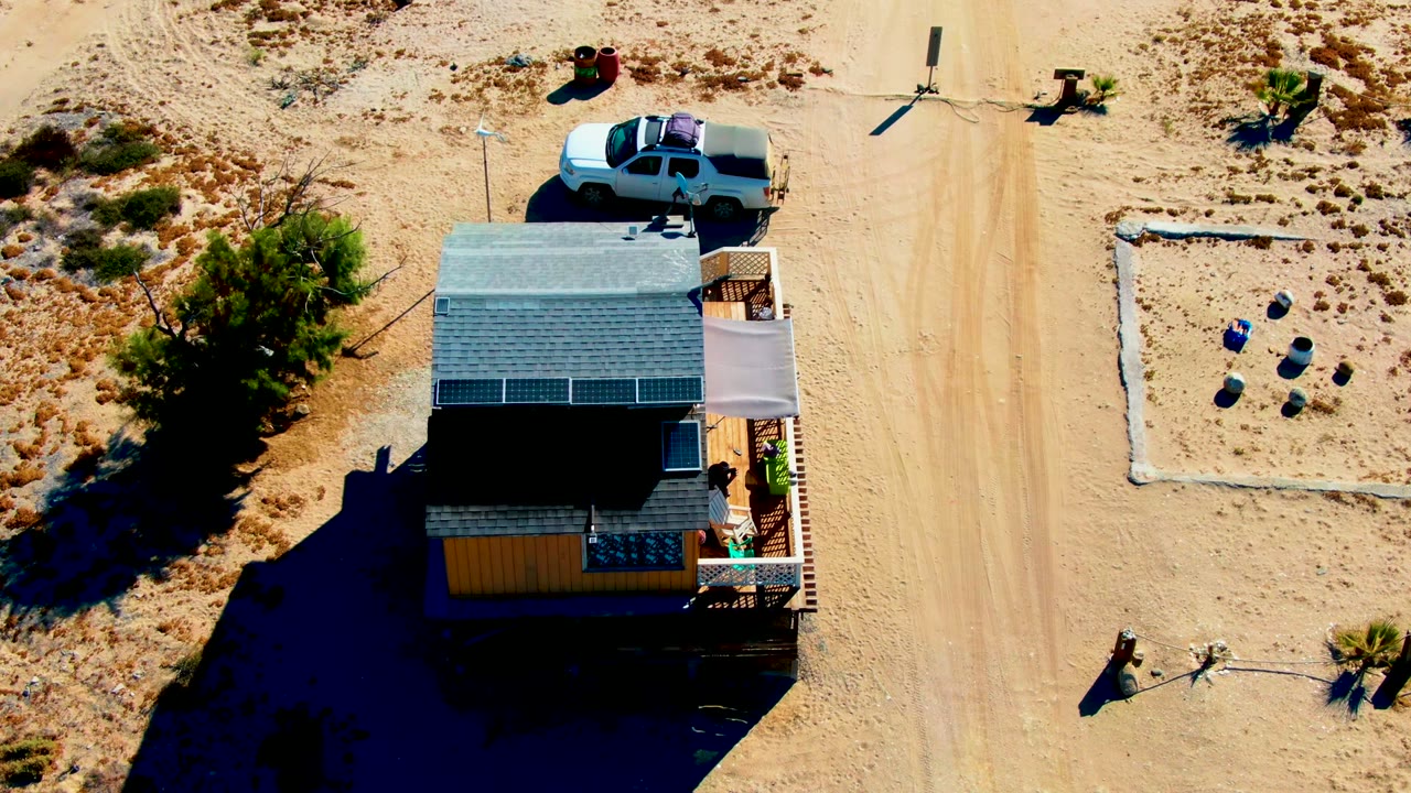 La Bocana, Baja California Sur aerial frame showing the white truck above the roofline of a small house with solar panels, a deck shade cover, sandy tracks, and no visible people.