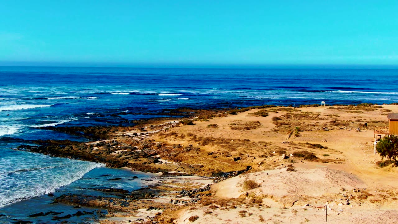 Opening frame of La Bocana, Baja California Sur shoreline with tan sand in front, dark rock shelves below, blue ocean bands beyond, and no visible people.