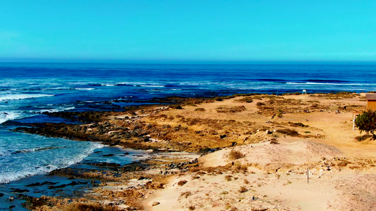 A wide La Bocana shoreline view shows dry sand and sparse scrub in the foreground, dark rocks and white surf in the midground, and no visible people.