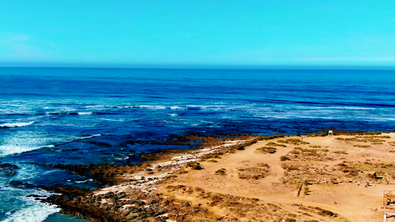 At La Bocana, Baja California Sur, a wide elevated view shows blue ocean, white surf lines, dark rocks, tan sand, sparse scrub, and no visible people.