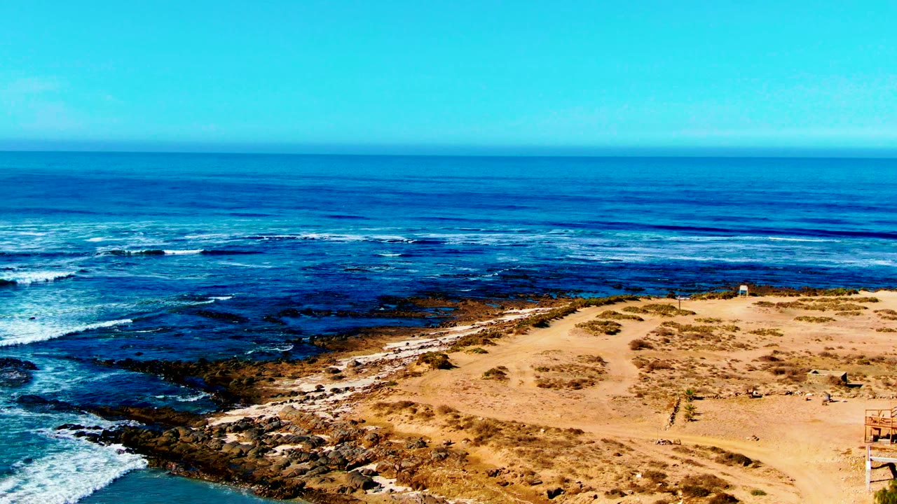 Ocean waves break around dark rocks beside a sandy coastal bluff.