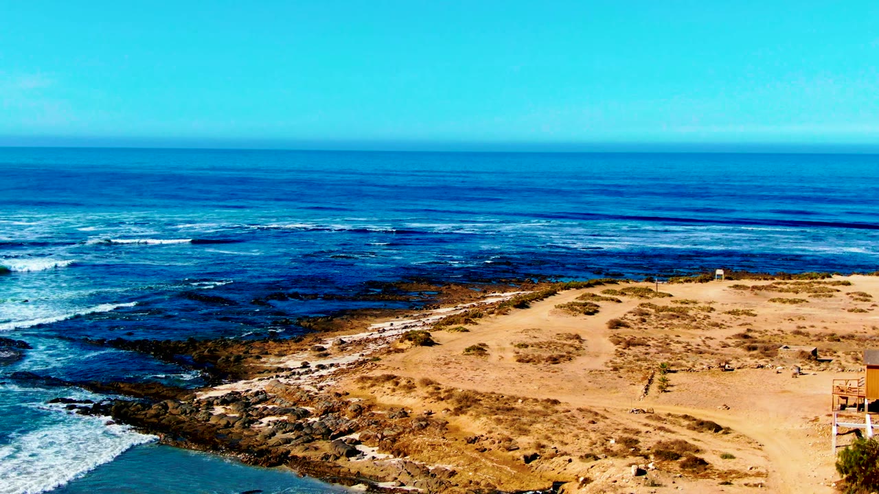 At La Bocana, Baja California Sur, the frame holds a broad ocean horizon above white wave bands, dark shoreline rocks, tan sand, scrub patches, and no visible people.