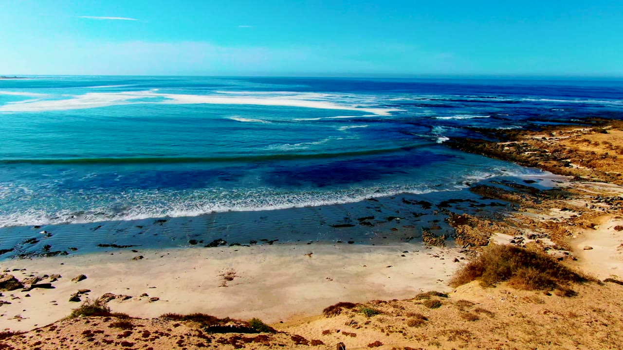 Aerial frame over La Bocana showing turquoise ocean, dark reef patches, pale sand, tan rocks at right, a clear blue sky, and no visible people at the opening moment.