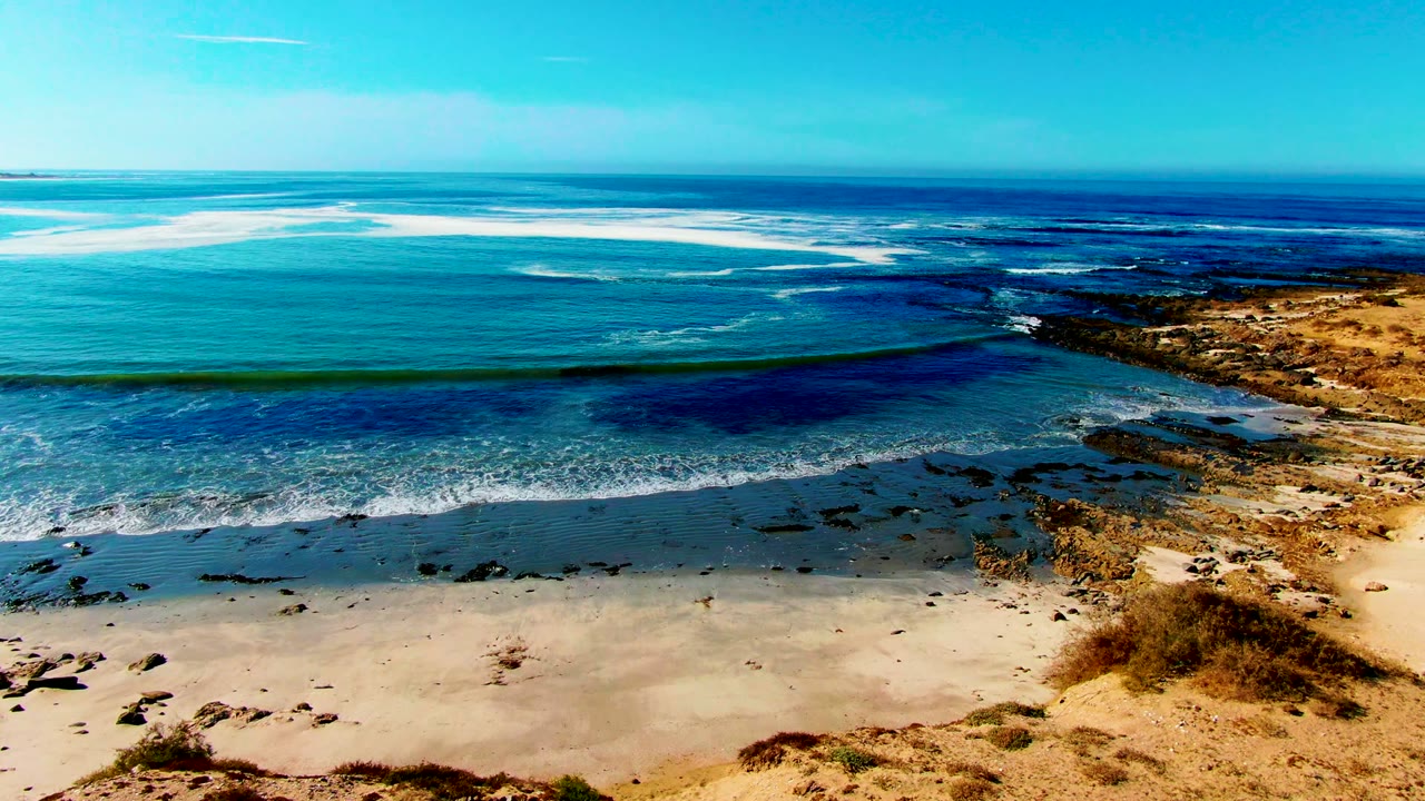 Blue-green surf spreads toward a sandy beach beside rocky coastline.