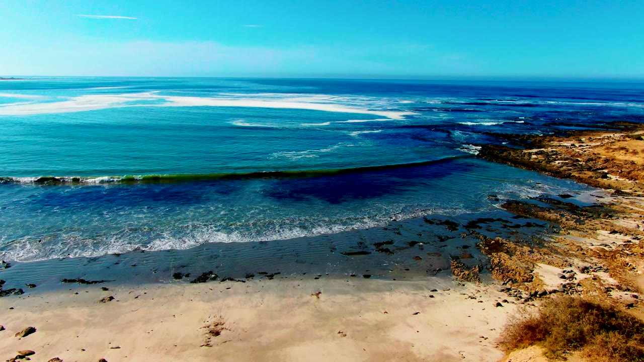 La Bocana shoreline seen from above as a dark swell band crosses shallow water, white foam touches the sand, rocks sit at the right edge, and no visible people appear.