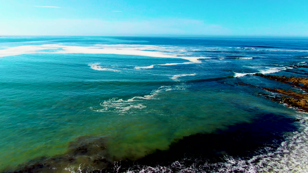 Aerial view over La Bocana, Baja California Sur, with turquoise water in the foreground, white surf over darker reef, rocky coast at right, clear sky, and no visible people.