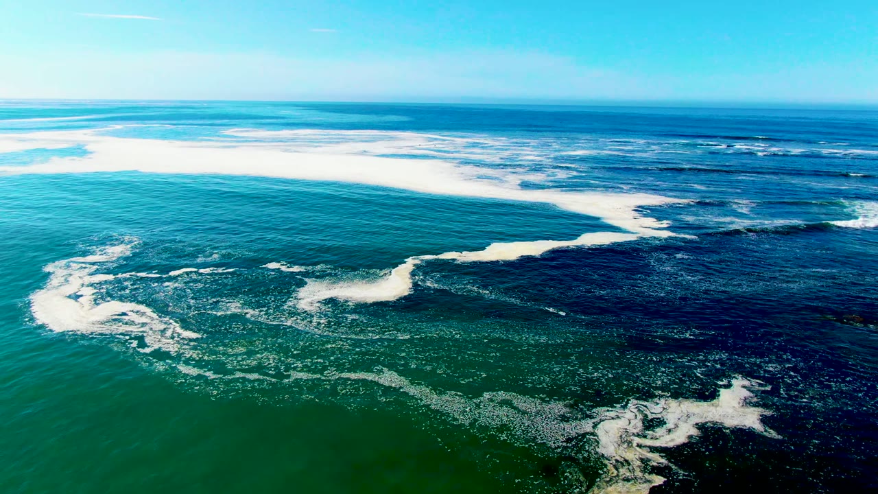 Aerial view over La Bocana, Baja California Sur, with white surf foam curving across blue-green ocean water and no visible people.