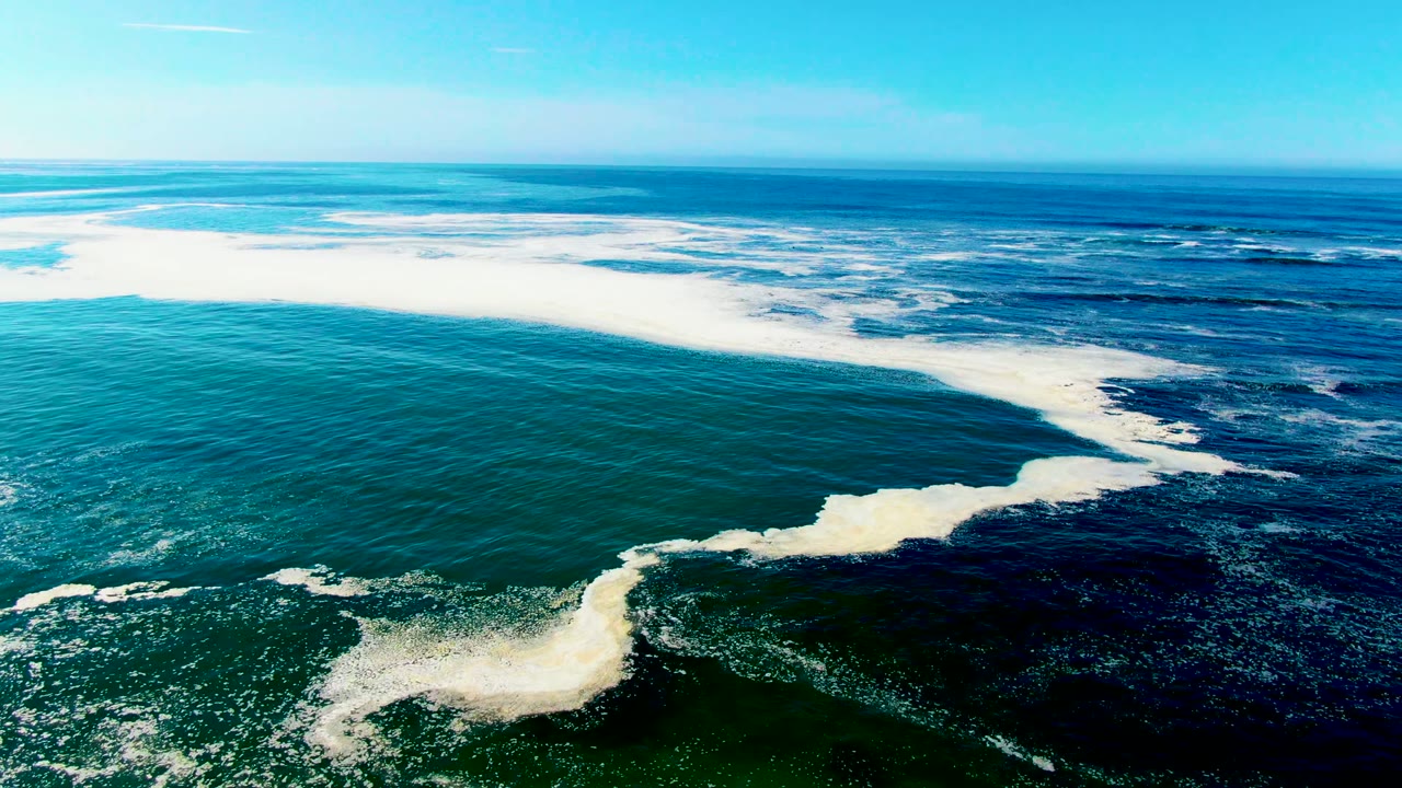 Aerial view over La Bocana with a wide white surf band stretching across the frame and deep blue water beyond, with no visible people.