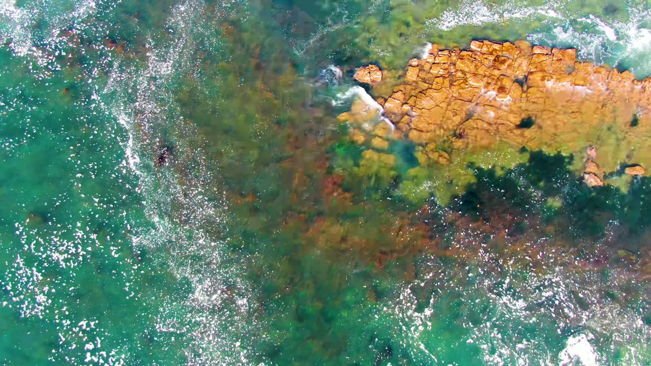 Overhead view in La Bocana, Baja California Sur, with clear turquoise water, ochre shoreline rocks, small white foam streaks, and no visible people.