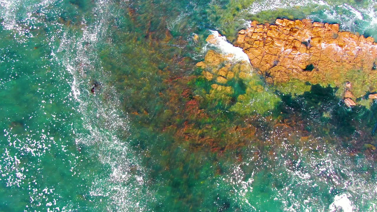 Clear green-blue water surrounds a rocky outcrop with foam along its upper edge.