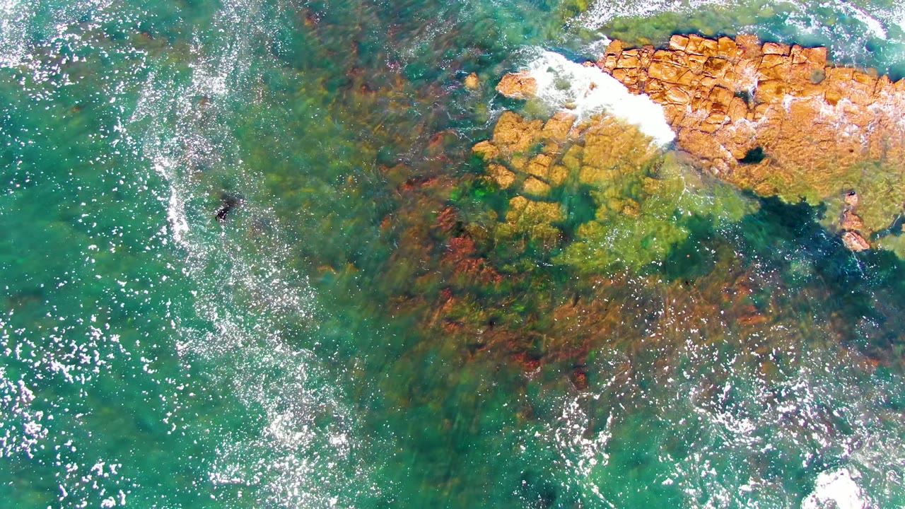 Overhead La Bocana footage shows a larger band of white foam breaking across the top-right rocks, with clear green water and no visible people.