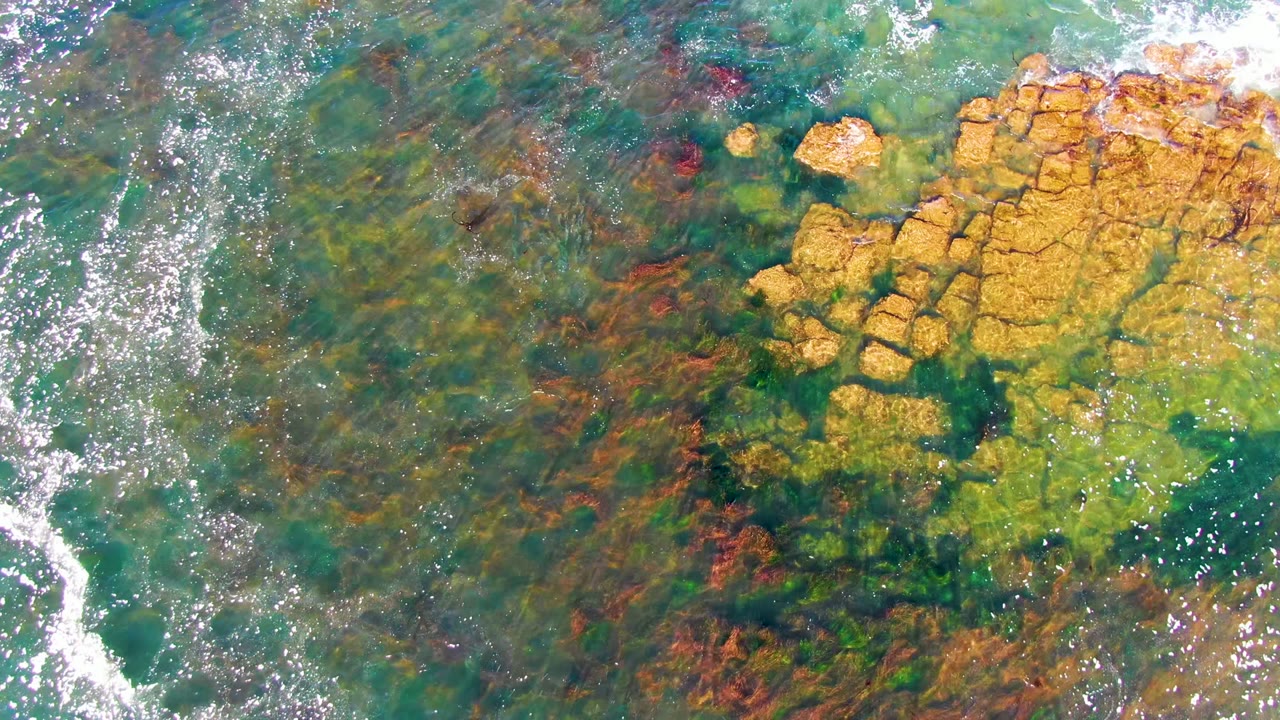 At two seconds, the overhead La Bocana frame shows brighter sun reflections across the water and the main yellow rock cluster near the upper right, with no visible people.