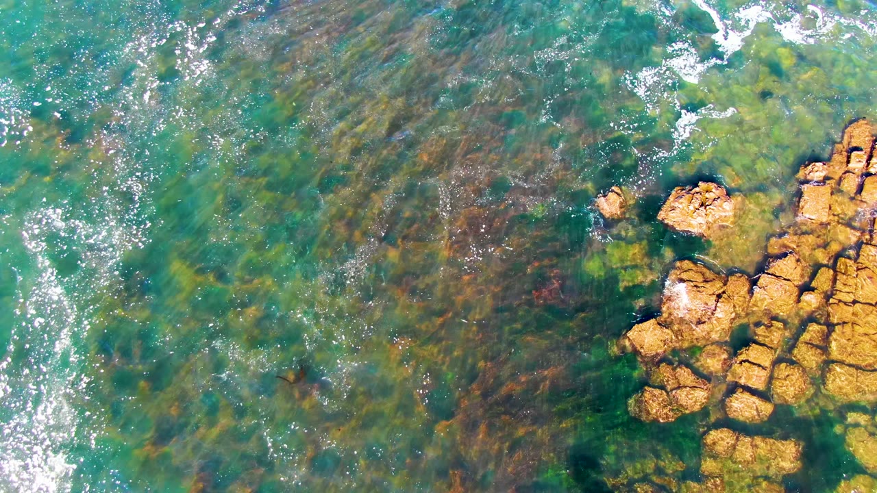 Aerial view over La Bocana shallow water where a larger rock formation enters from the right and darker seabed patches show beneath the surface, with no visible people.