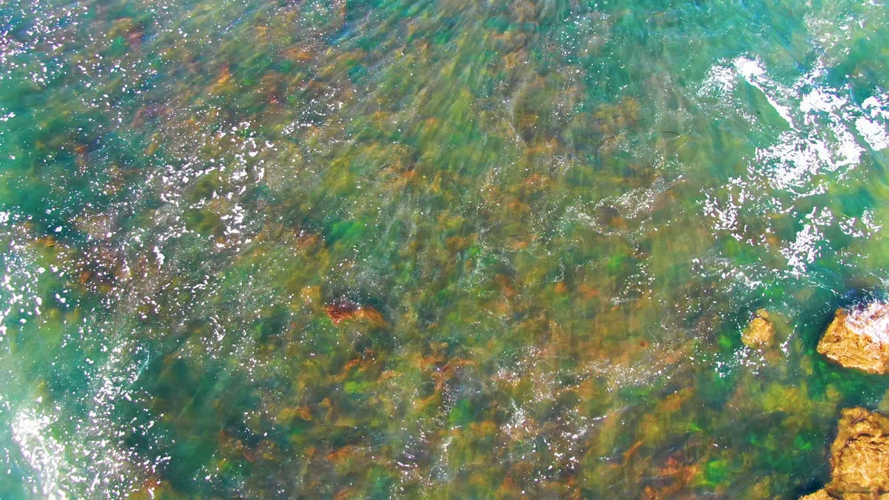 Top-down view of turquoise and green shallow water over rocks in La Bocana, Baja California Sur, with scattered white foam and no visible people.