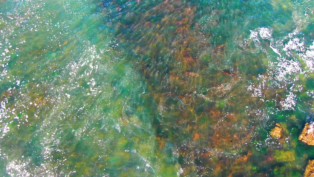 Aerial-looking top-down frame of shallow water at La Bocana, Baja California Sur, with tan rocks near the lower edge, streaked green seabed, and no visible people.