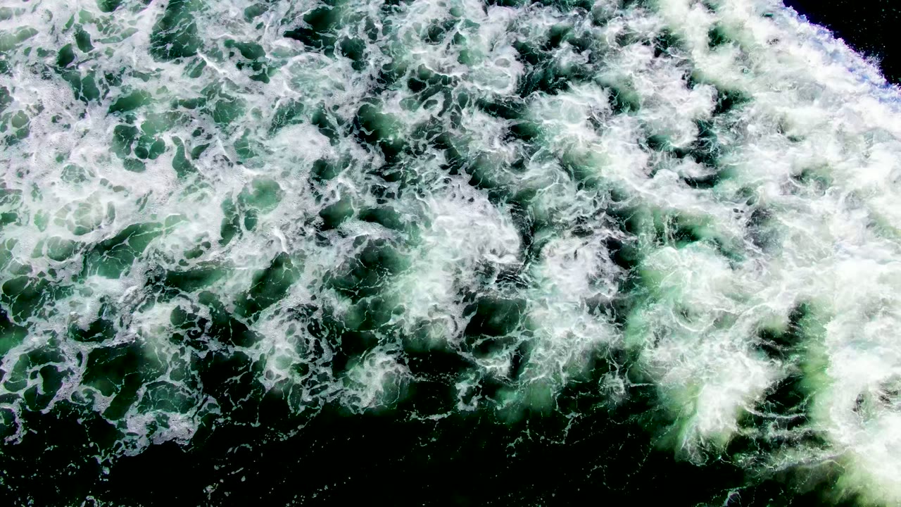 Broken white froth patterns cover green seawater in La Bocana, Baja California Sur, with small darker channels opening through the foam and no visible people.