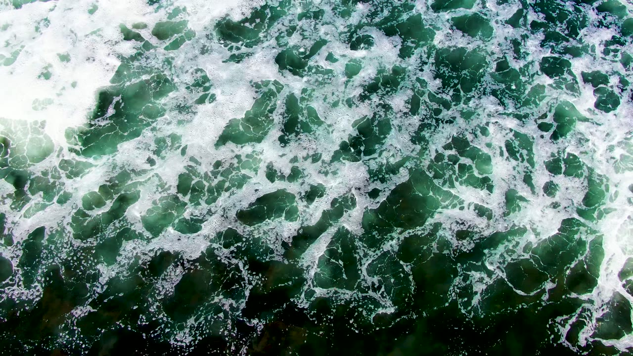 Top-down view of white surf foam spreading over green ocean water in La Bocana, Baja California Sur, with darker patches below and no visible people.