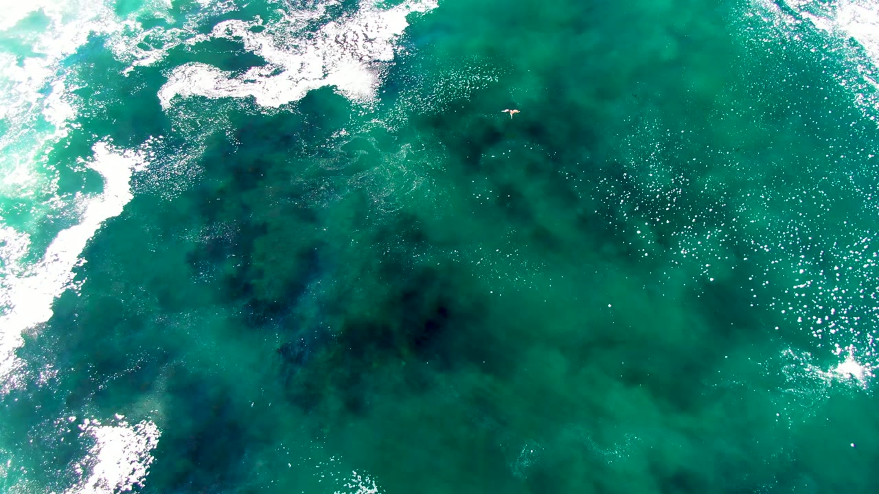 Aerial frame over turquoise water at La Bocana, Baja California Sur, with white surf entering from the upper left and dark submerged patches below, no visible people.