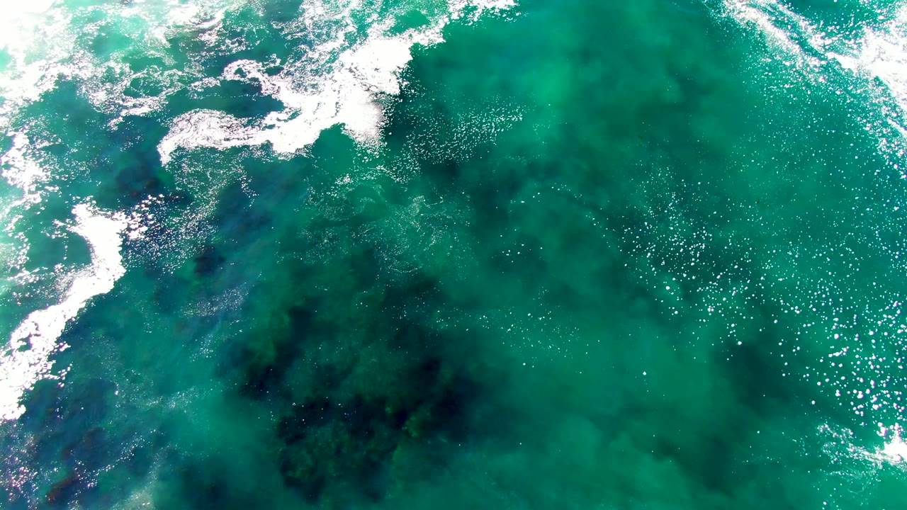 Top-down La Bocana ocean frame with a curved white foam break, turquoise water, darker patches beneath the surface, and a tiny pale speck too small to identify as a person.