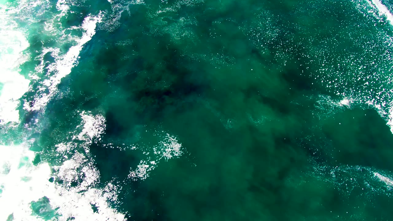 Top-down view of turquoise ocean water at La Bocana, Baja California Sur, with white foam entering from the left edge and no visible people.