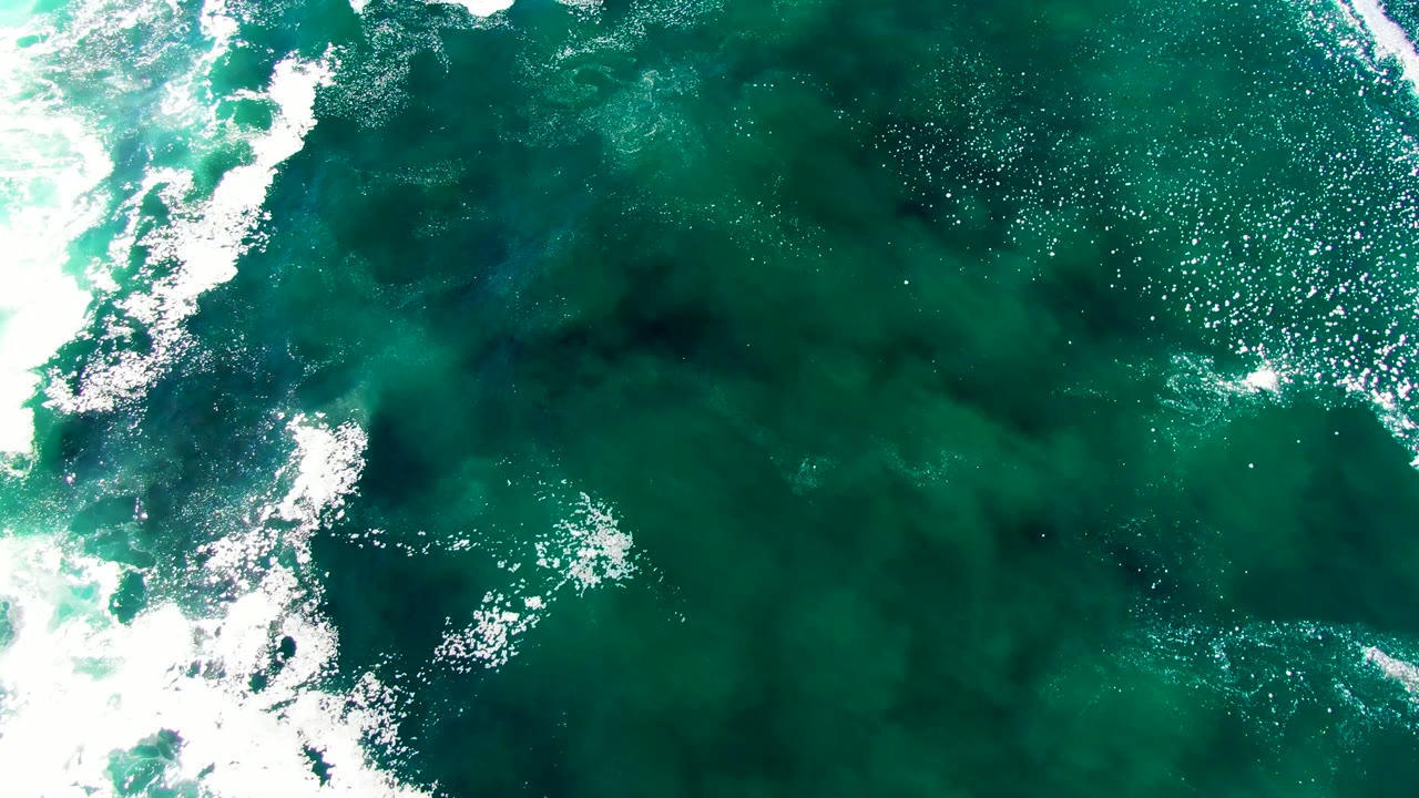Overhead La Bocana sea surface with white foam crossing the left third, scattered sunlight highlights on turquoise water, and no visible people.