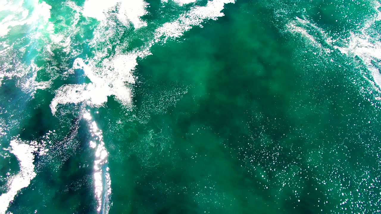 Top-down aerial view of turquoise ocean water at La Bocana with white foam entering from the upper edge, bright sun reflections, darker green patches, and no visible people.