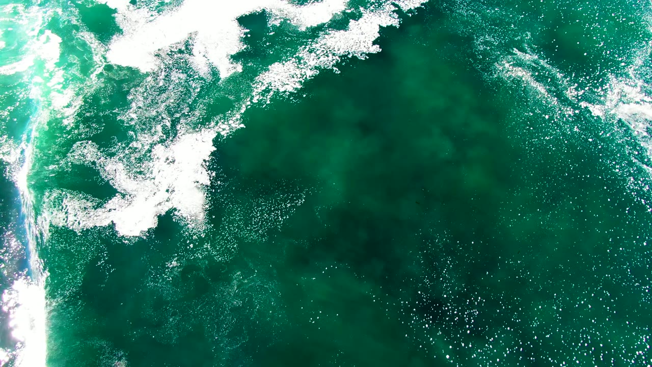 Top-down La Bocana sea surface with a bright foamy break along the upper left, a vertical strip of reflected light near center right, dark green water, and no visible people.