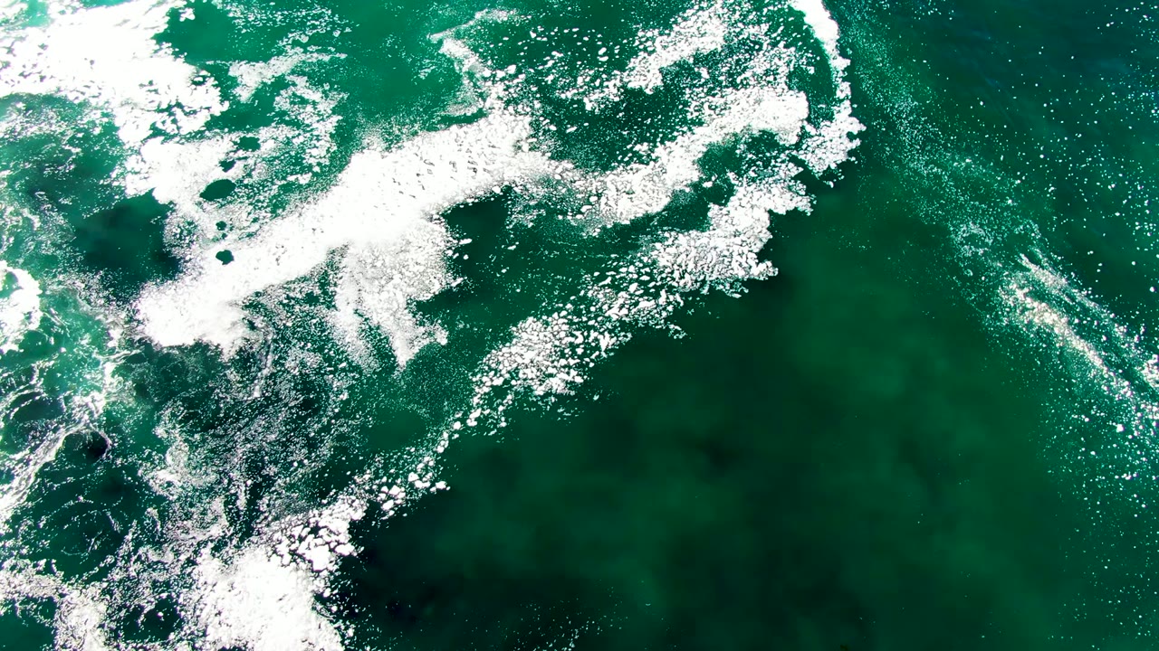 Aerial overhead frame from La Bocana showing white foam drifting through green ocean water, with a darker patch near the lower left and no visible people.