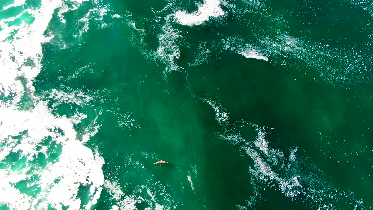 Aerial view over green ocean water at La Bocana, Baja California Sur, with white foam on the left and a small orange surfboard visible near the lower left, no visible people.
