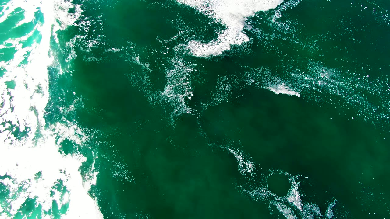 Aerial view of darker green ocean at La Bocana with scattered white foam and a broad curved patch of whitewater near the lower center, no visible people.