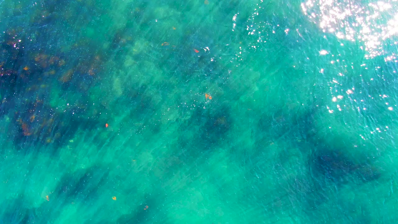 Overhead view of turquoise ocean water in La Bocana, Baja California Sur, with darker blue-green patches, bright surface glare, and no visible people.